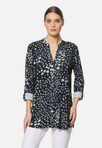 Schwarze Bluse mit hellblauen und weißen Punkten, mit Kragen und langen Ärmeln, die an den Bündchen hochgerollt sind, in einem lockeren, fließenden Design.