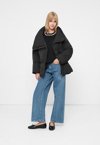 Γυναίκα που στέκεται με το χέρι στην τσέπη, φορώντας μια υπερμεγέθη μαύρη πούφα, μαύρο πουλόβερ, φαρδύ τζιν σε μπλε απόχρωση και μαύρα μπαλαρίνα.