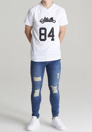 Osoba ubrana w białą koszulę z krótkim rękawem z napisem „Siksilk 84”, podniszczone niebieskie obcisłe dżinsy i białe trampki na jednolitym tle.