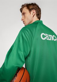Zelena atletska jakna glatke teksture, s riječju "CELTICS" ispisanom velikim bijelim slovima na leđima. Košarka se drži u ruci.