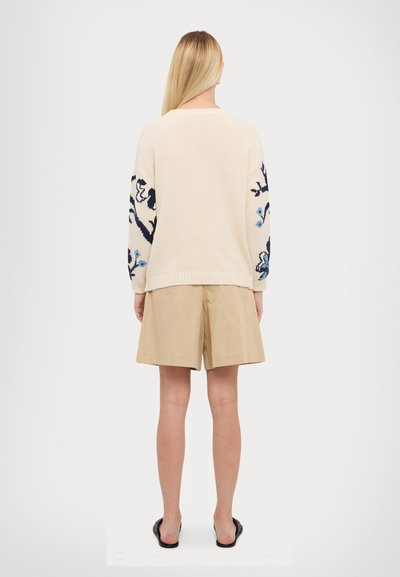 Suéter de punto crema con bordado floral azul en las mangas, combinado con pantalones cortos beige. La tela es suave y tiene un corte relajado.