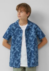 Kurzärmliges Hemd aus blauer Baumwolle mit einem Vogel-Muster, mit Kragen und lockerer Passform, über einem weißen T-Shirt getragen.