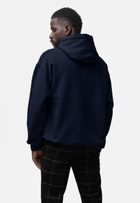 Felpa con cappuccio blu scuro dalla vestibilità comoda e tessuto strutturato, vista da dietro. Pantaloni neri a quadretti con linee bianche.