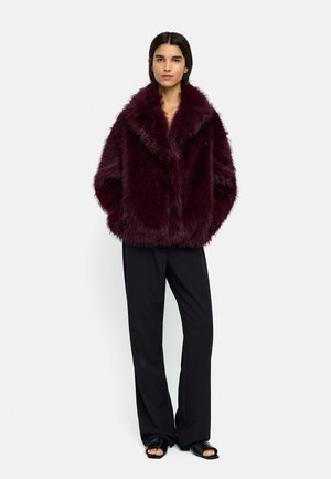 Kobieta stoi z rękami w kieszeniach, ubrana w ciemno bordowy płaszcz z futerka sztucznego, czarne szerokie spodnie i czarne buty z odkrytymi palcami.