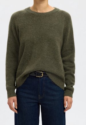 Persoon draagt een donkergroen, pluizige trui die in een donkerblauwe spijkerbroek is gestopt, met een zwarte riem met een gouden gesp, tegen een effen lichte achtergrond.