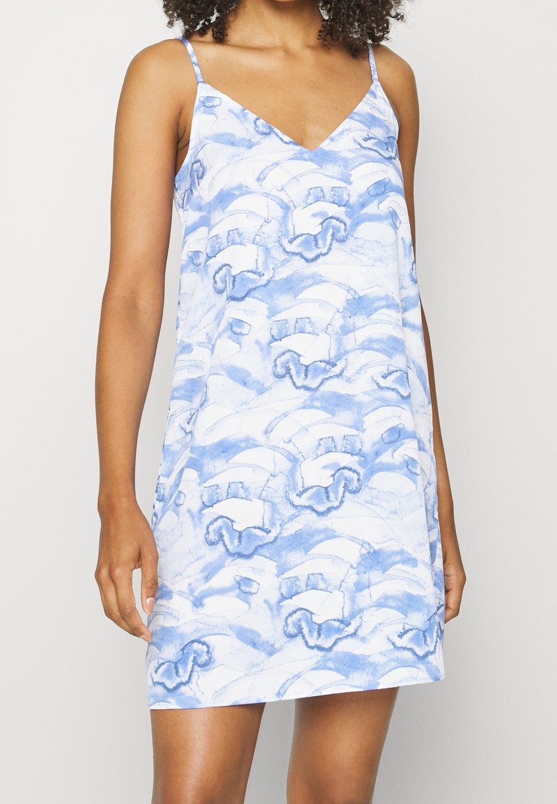 Femme portant une robe sans manches bleu et blanche à motif, à col en V avec de fines bretelles, montrée des épaules jusqu'à la mi-cuisse.
