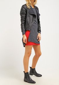 Veste en cuir noir, robe grise avec un ourlet rouge et bottines noires. La tenue combine des éléments lisses et texturés, mettant en avant le contraste.