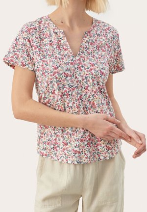 Vrouw draagt een korte mouwen bloes met roze, rode en gele bloemen, gecombineerd met lichtbeige broek, handen zachtjes voor zich gevouwen.