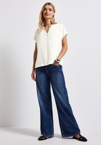 Leichte, kurzärmlige Bluse in Creme mit V-Ausschnitt, kombiniert mit lockeren, dunkelblauen weit geschnittenen Jeans und schwarzen geflochtenen Ballerinas.