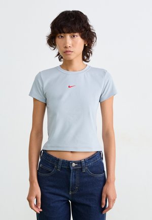 Młoda osoba z kręconymi ciemnymi włosami w jasnoszarym, krótkim t-shircie Nike i ciemnoniebieskich dżinsach, stojąca na tle białej ściany.