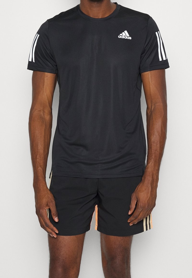 Homme portant un t-shirt noir à manches courtes Adidas avec des bandes blanches sur les manches et un short noir assorti avec des bandes latérales beiges.
