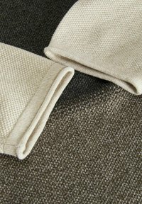 Det stickade tyget har två beige element med texturerade mönster, placerade mot en mörkgrön bakgrund. Kanterna är prydligt vikta.