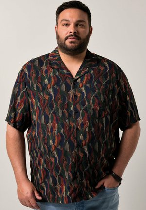 Mann mit kurzen dunklen Haaren und Bart, trägt ein bunt gemustertes kurzärmliges Hemd und hellblaue Jeans, steht mit einer Hand in der Tasche.
