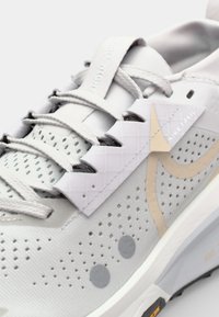 Hellgraue Sneaker mit einem atmungsaktiven Mesh-Obermaterial, perforierter Textur, reflektierenden Akzenten und einer flexiblen Sohle mit Profildetails.