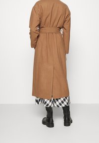 Persona che indossa un cappotto color cammello con cintura sopra una gonna a scacchi bianco e nero, con stivali di pelle nera, in piedi contro uno sfondo bianco.