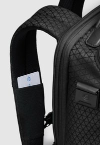 Zwarte rugzak met een gestructureerd diamantpatroon. Voorzien van een gevoerde schouderband en een opbergvak met een kaart erin. Ritsvak zichtbaar.