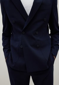 Giacca blu navy doppiopetto con texture liscia, due bottoni e una tasca sul petto. I pantaloni hanno una vestibilità sartoriale.