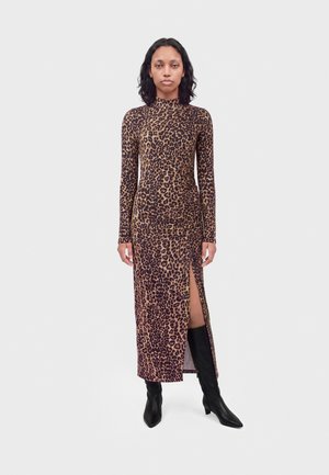 Femme debout portant une robe à manches longues imprimé léopard avec une fente haute et des bottes noires montantes jusqu'aux genoux sur un fond uni.