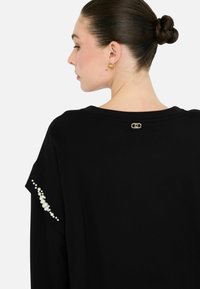 Donna con i capelli raccolti in uno chignon, che indossa orecchini d'oro e una maglia nera con dettagli in perle sulle maniche e un piccolo logo dorato sulla scollatura posteriore.