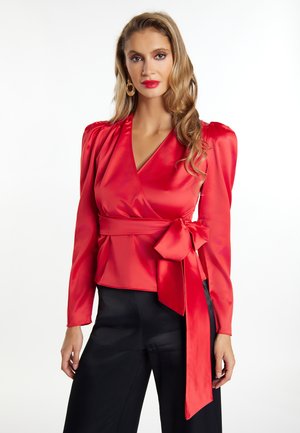 Top avvolgente in satin rosso con maniche a soffice, scollatura profonda a V e vita legata. Abbinato a pantaloni neri con una texture liscia.