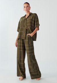 Maslinasto zelena dvodijelna odjeća koja se sastoji od košulje s kratkim rukavima na dugme i hlača širokih nogavica, oboje s bijelim geometrijskim uzorcima.