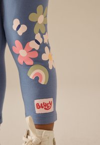 Leggings azules con apliques florales y de mariposas en rosa, verde y blanco, con un diseño de arcoíris y un parche con el logo "BLUEY".