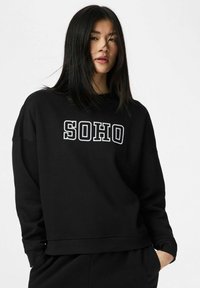 Jauna moteris su tiesiais juodais plaukais, dėvinti juodą džemperį, ant kurio užrašyta „SOHO“, stovinti su rankomis kišenėse prieš balto fono foną.