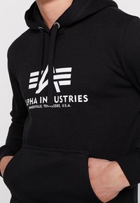 Czarna bluza z kapturem z białym nadrukiem logo, na którym widnieje napis "ALPHA INDUSTRIES" oraz graficzny motyw dwóch poziomych linii nad tekstem.