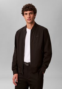 Veste bomber noire à texture lisse, fermeture éclair sur le devant, poignets et ourlet côtelés, associée à un t-shirt blanc à col rond et un pantalon noir.