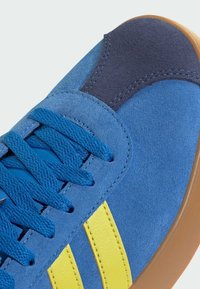 Sneaker blu scamosciato con strisce gialle, dotato di una parte superiore testurizzata, lacci tondi e una suola in gomma naturale. I dettagli includono pannelli blu scuro.