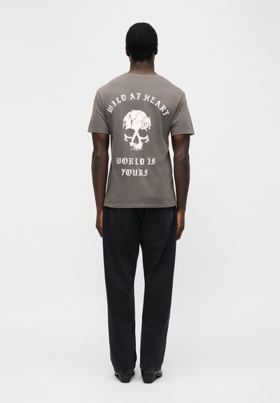 Άντρας που φορά γκρι μπλουζάκι με λευκό γραφικό κρανίο και κείμενο «WILD AT HEART» και «WORLD IS YOURS» στην πλάτη, μαύρο παντελόνι, μαύρα παπούτσια, στέκεται με την πλάτη προς τα εμπρός.