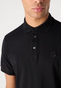 Črn polo majica z teksturirano tkanino, tremi gumbi in vezenim logotipom. Ima klasičen ovratnik in kratke rokave.