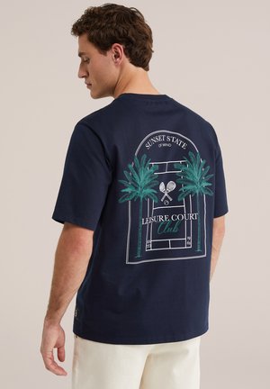 Muškarac nosi tamnoplavu majicu s grafikom dvije palme, ukriženih teniskih reketa i tekstom "Sunset State of Mind Leisure Court Club" na leđima.
