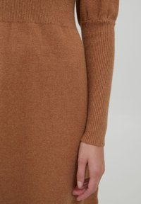 Abito lavorato a maglia marrone con una texture a costine sulle maniche. Il tessuto appare morbido e leggero, con una silhouette aderente.