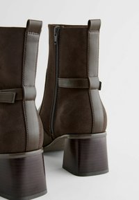 Bottines en suède marron avec un talon carré, une fermeture éclair sur le côté et des accents en cuir contrastant à l'arrière et sur les côtés. Fini mat.