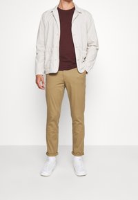 Ljusbeige jacka över en vinröd t-shirt, tillsammans med kaki chinos och vita sneakers. Jackan har en avslappnad passform och synliga knappar.