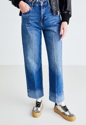 Jeans in denim blu a gamba larga con dettagli sbiaditi. Abbinati a sneakers con motivo bianco e nero e una giacca di pelle nera sbottonata.