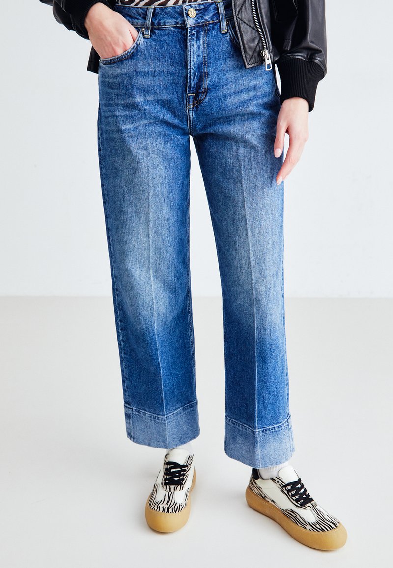 Jeans de mezclilla azul de pierna ancha con detalles desgastados. Combinados con zapatillas de patrón blanco y negro y una chaqueta de cuero negra desabotonada.