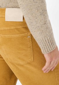 Gele corduroy broek met verticale ribbels, voorzien van een leren label op de tailleband en een klein metalen merkembleem op de achterzak.