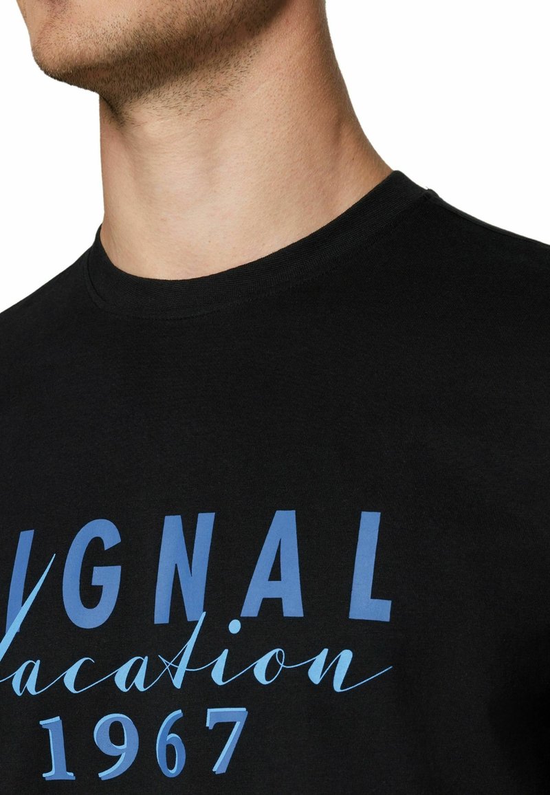 Κοντινό πλάνο ενός ατόμου που φοράει μαύρο T-shirt με μπλε κείμενο που γράφει "ORIGINAL vacation 1967" στο στήθος.