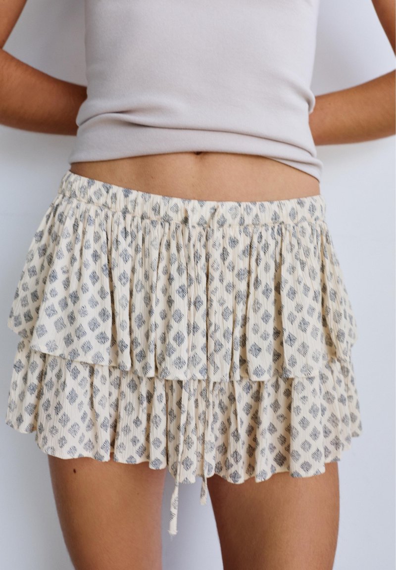 Femme portant une mini-jupe beige à volants à motifs et un top gris clair sans manches, debout avec les bras derrière le dos contre un mur uni.