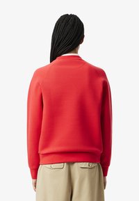 Felpa rossa con colletto rotondo, maniche raglan e polsini a coste. Il tessuto appare liscio con una morbida texture, abbinata a pantaloni beige.