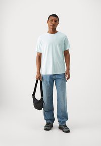 Camiseta de manga corta azul claro, corte relajado, combinada con unos vaqueros holgados azul claro. Sosteniendo un pequeño bolso cruzado negro, lleva zapatillas gris.