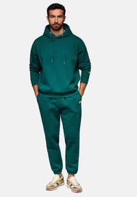 Turkos hoodie med dragsko, kombinerad med matchande joggingbyxor. Båda i mjukt tyg. Sneakers med beige och gula accenter.