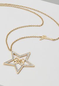 Collier chaîne en or avec pendentif en forme d'étoile bordé de cristaux et logo "G" entrelacé à l'intérieur, avec une étiquette "Guess" sur la chaîne.