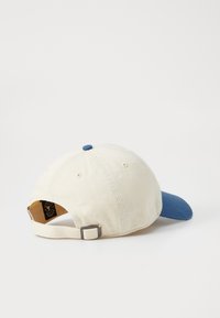 Katoenen pet met een crèmekleurige kroon en een blauwe klep. Heeft een verstelbare band en een klein topknopje. De buitenkant heeft een gladde textuur.