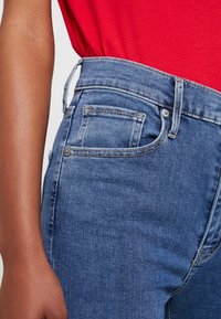 Jeans de mezclilla azul de cintura alta, con una textura suave, diseño de cinco bolsillos, lazos para cinturón y detalles de costura en contraste.