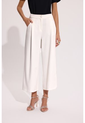 Kobieta ubrana w białe spodnie z wysokim stanem i szerokimi nogawkami z przednimi plisami, czarny top oraz srebrne, paskowe sandały na wysokim obcasie, stojąca z jedną ręką w kieszeni.