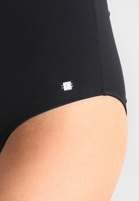 Maillot de bain noir avec une texture lisse et extensible, doté d'un petit accent de logo argenté sur le côté et de coutures nettes le long des bords.