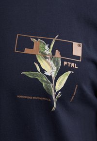 Marine katoenen t-shirt met een botanische grafische print van groene bladeren en witte bloemen, geaccentueerd met gouden tekst en een rechthoekig logo.